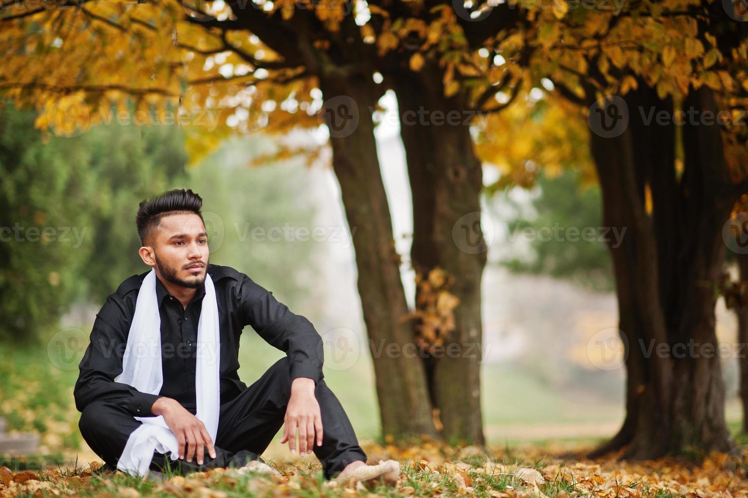 hombre indio con estilo en ropa tradicional negra con bufanda blanca posada al aire libre contra el árbol de hojas amarillas de otoño. foto