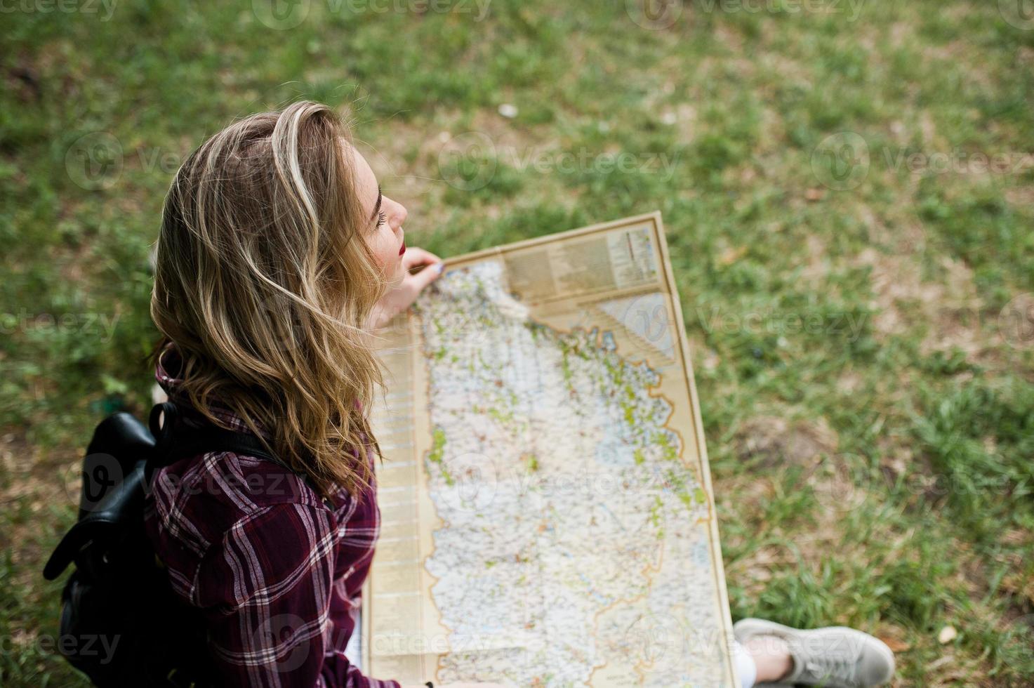 Portrait of a positive young gorgeous blonde sitting on the ground with a map in her hands in the forest. photo