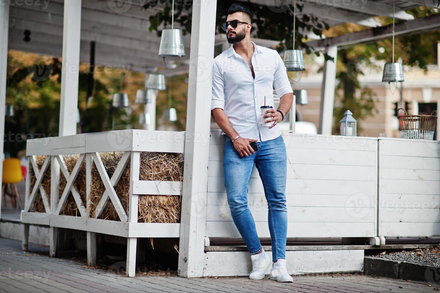 Stylish tall arabian man model in white shirt, jeans and sunglasses posed at street of city. Beard attractive arab guy with cup of coffee against outdoor cafe. photo