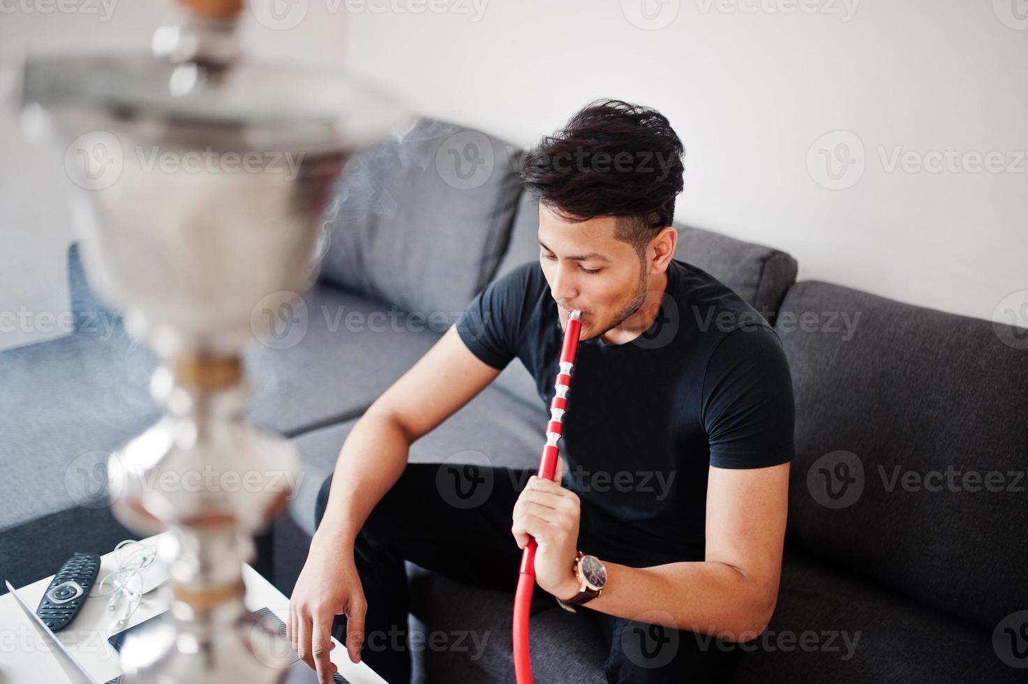 hombre indio guapo y de moda en negro sentado en la habitación, fumando narguile y trabajando en la computadora portátil. foto