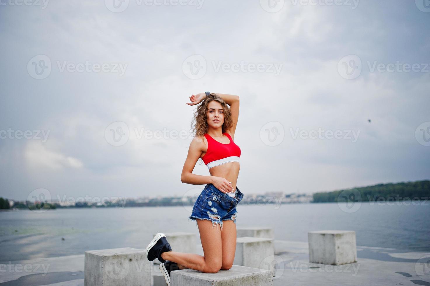 Sexy curly model girl in red top, jeans denim shorts and sneakers posed at stone cubes against cloudy sky and lake. photo
