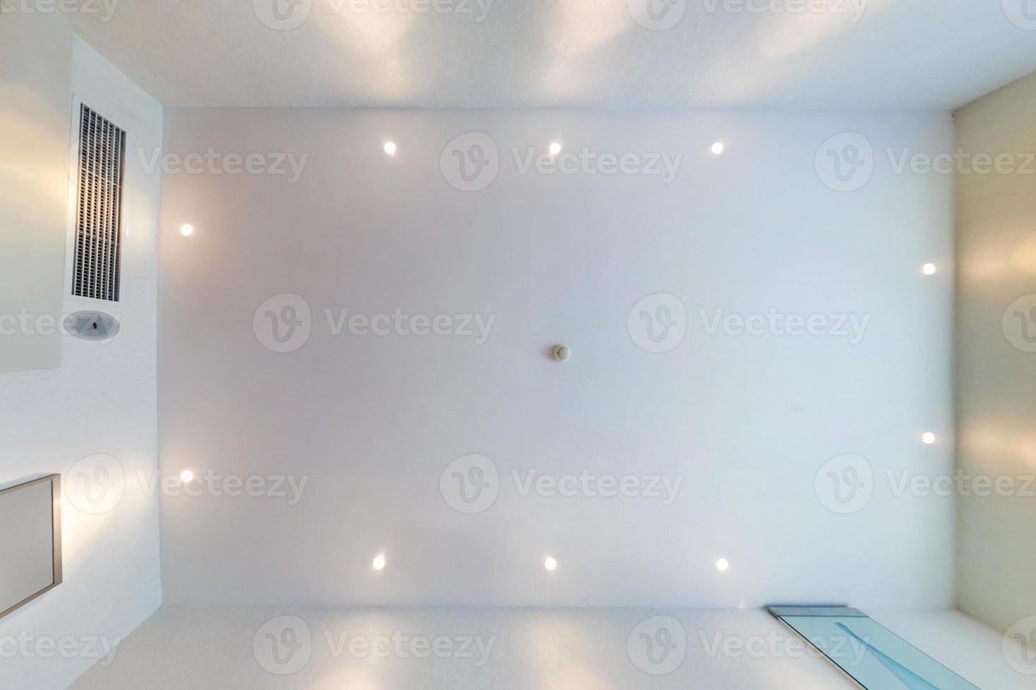 look up on suspended ceiling with halogen spots lamps and drywall construction with fire alarm sensor in empty room in apartment or house. Stretch ceiling white and complex shape. photo
