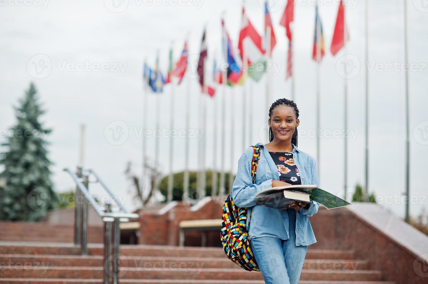 African student female posed with backpack and school items on yard of university, against flags of different countries. photo