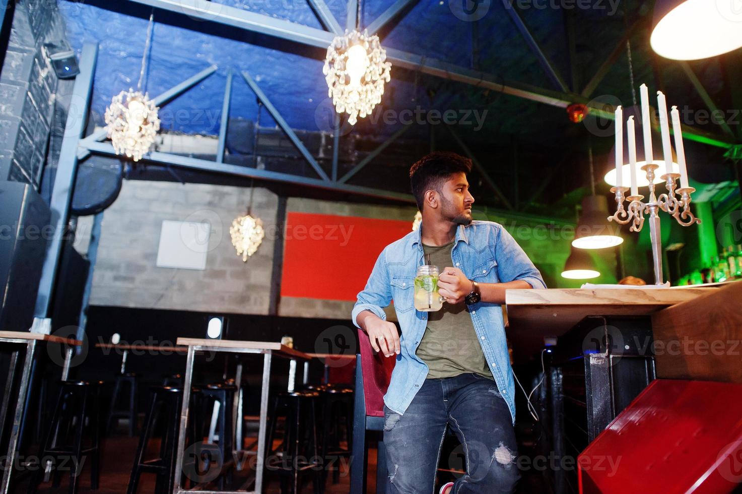 Portrait of handsome successful bearded south asian, young indian freelancer in blue jeans shirt sitting in night club against bar counter with cocktail and having a rest. photo