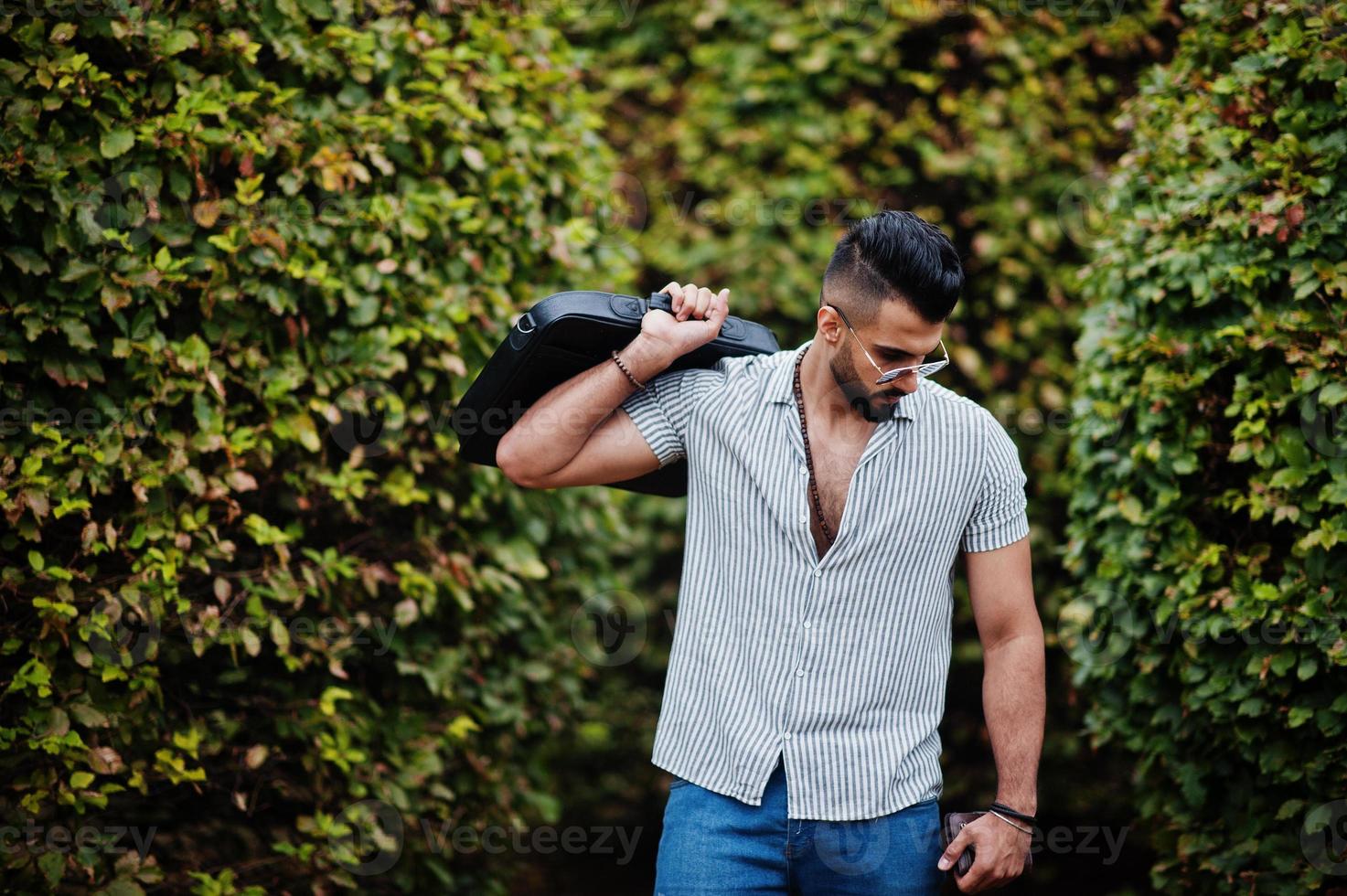 el hombre de barba árabe alto de moda usa camisa, jeans y gafas de sol posados en el parque y sostiene la caja de la bolsa. foto