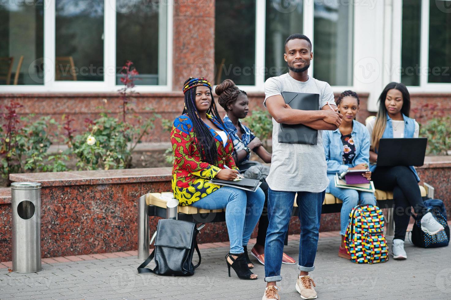 grupo de cinco estudiantes universitarios africanos que pasan tiempo juntos en el campus en el patio de la universidad. amigos afro negros que estudian en un banco con artículos escolares, computadoras portátiles. foto