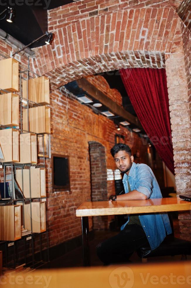 Portrait of handsome successful bearded south asian, young indian freelancer in blue jeans shirt sitting in cafe and having a rest. photo