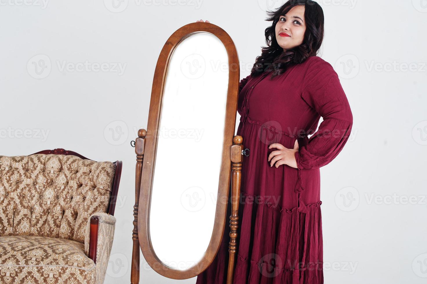 atractiva mujer del sur de asia con un vestido rojo profundo posó en el estudio sobre fondo blanco contra el espejo y la silla. foto