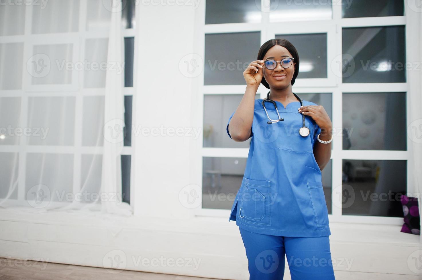 Portrait of happy female african american young doctor pediatrician in blue uniform coat and stethoscope against window in hospital. Healthcare, medical, medicine specialist - concept. photo