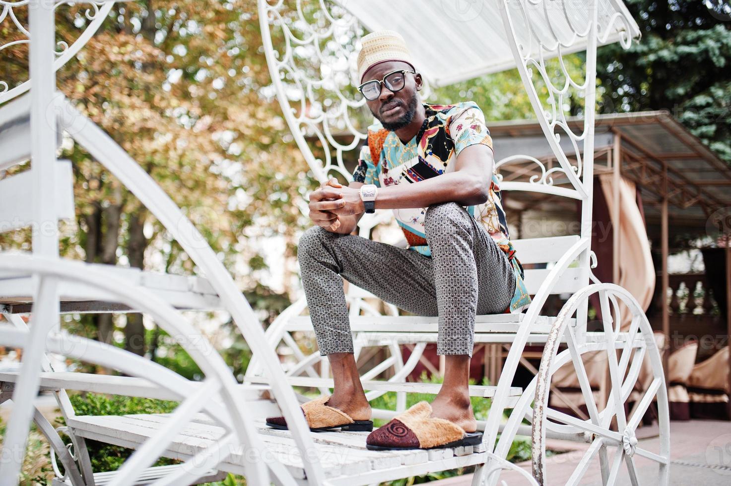 Handsome afro american man wearing traditional clothes, cap and eyeglasses sitting at white carriage. photo