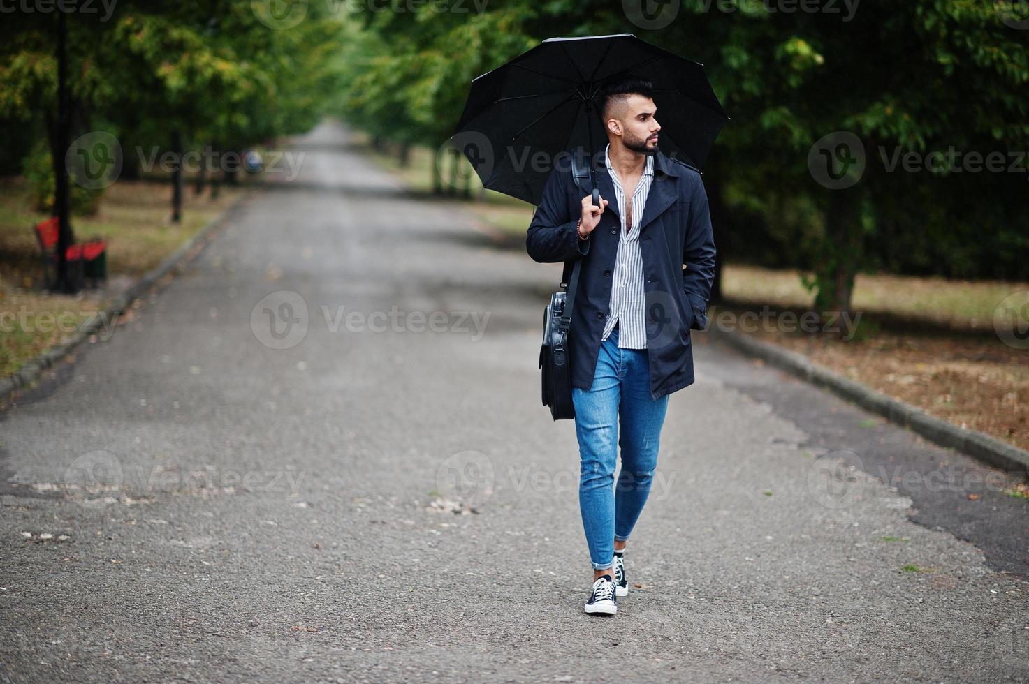 hombre de barba árabe alto de moda vestido con abrigo negro con paraguas y estuche posado en el día de la lluvia. foto