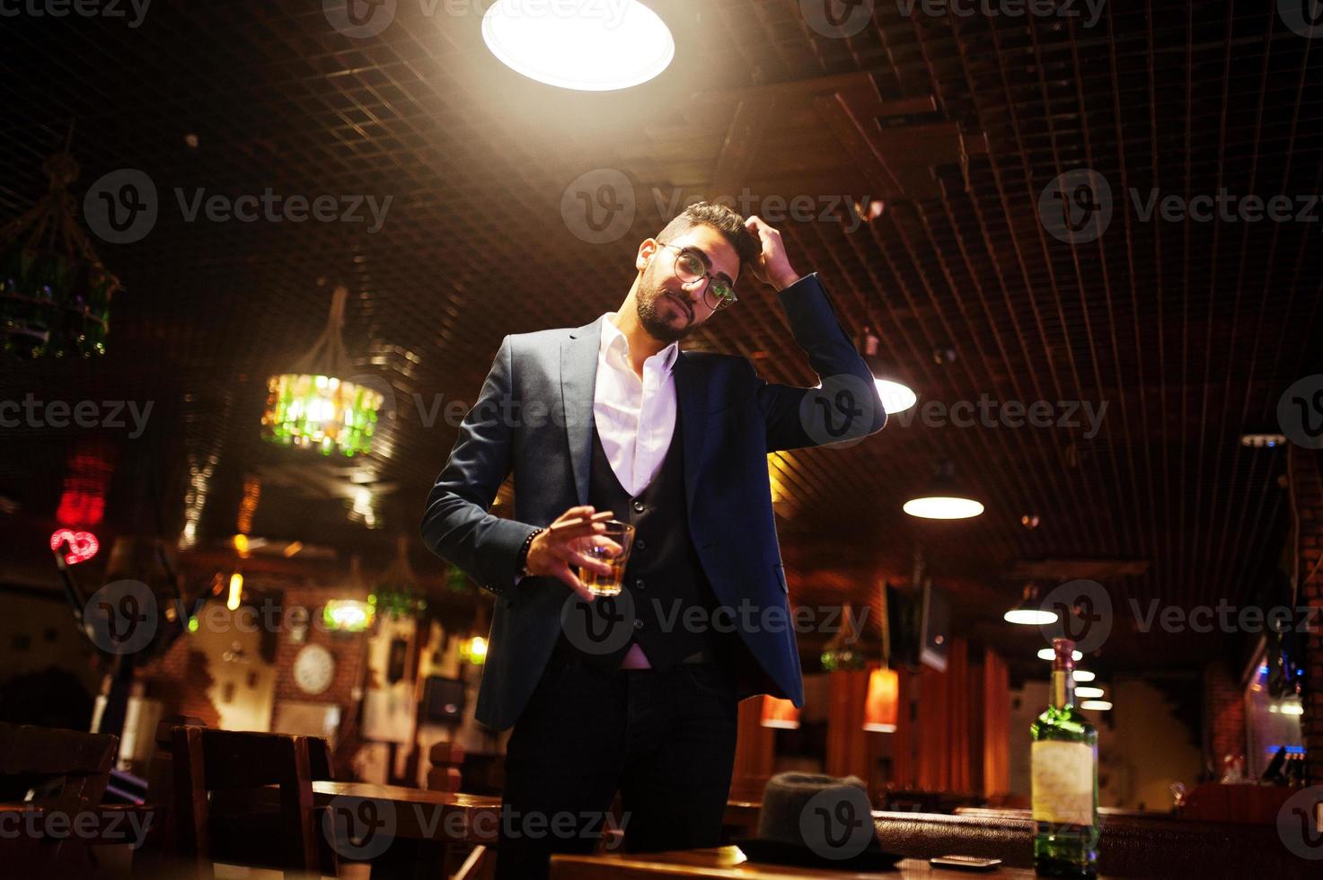 apuesto hombre árabe bien vestido con vaso de whisky y cigarro posado en el pub. foto