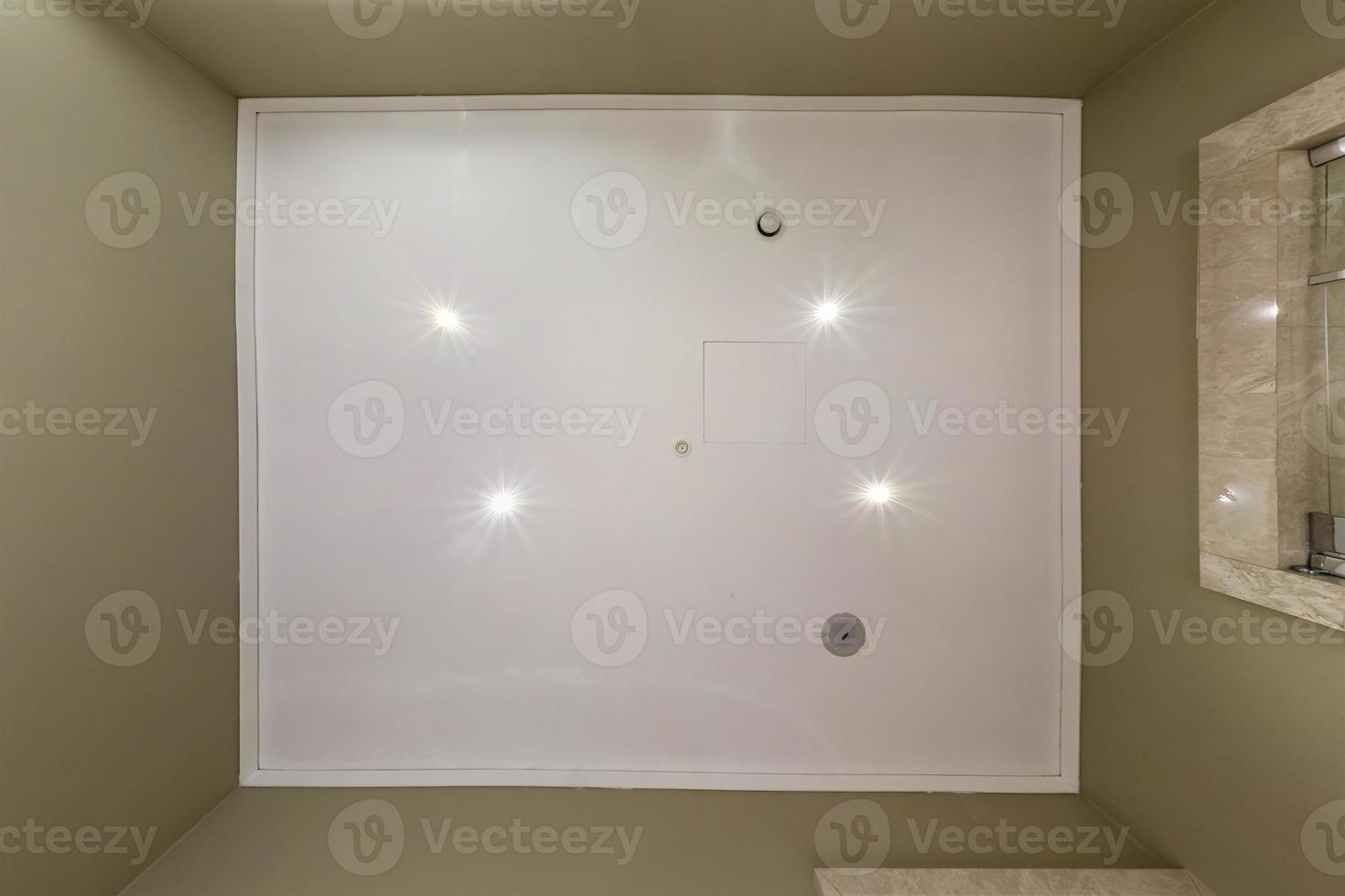 look up on suspended ceiling with halogen spots lamps and drywall construction with fire alarm sensor in empty room in apartment or house. Stretch ceiling white and complex shape. photo