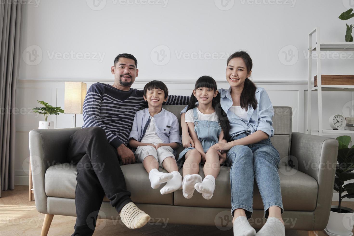 Portrait of Asian Thai family, adult dad, mum with little children happiness home living together, looking at camera, leisure on sofa in white room, lovely weekend and wellbeing domestic lifestyle. photo