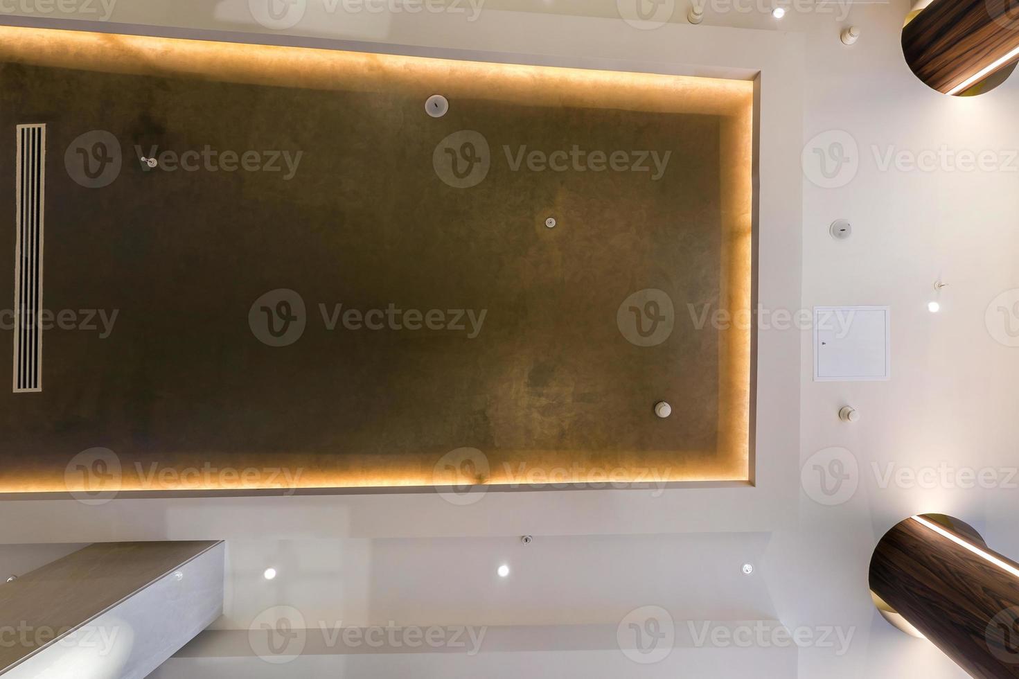 look up on suspended ceiling with halogen spots lamps and drywall construction with fire alarm sensor in empty room in apartment or house. Stretch ceiling white and complex shape. photo