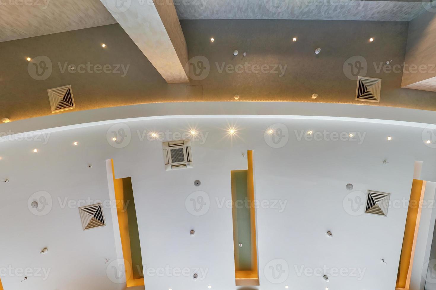 look up on suspended ceiling with halogen spots lamps and drywall construction with fire alarm sensor in empty room in apartment or house. Stretch ceiling white and complex shape. photo