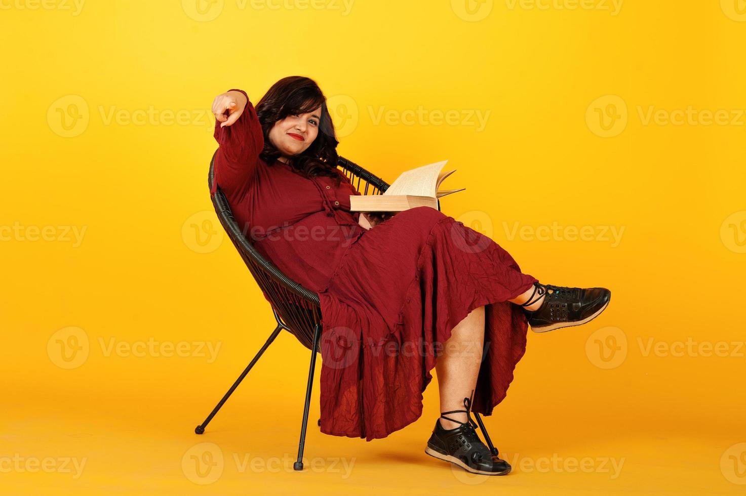 Attractive south asian woman in deep red gown dress posed at studio on yellow background sitting on chair with book. photo
