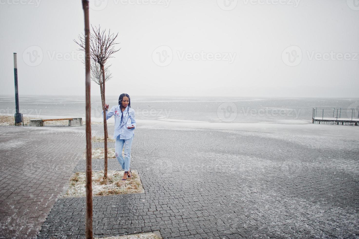 Stylish african american girl with dreads holding mobile phone at hand, outdoor on pier against frozen lake at snowy weather. photo