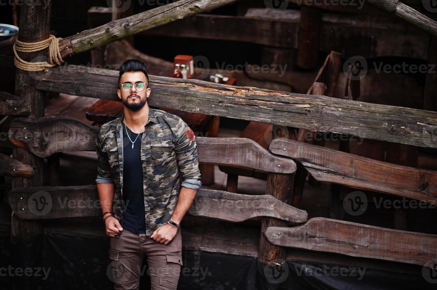 Impresionante hermoso hombre macho alto de barba árabe con gafas y chaqueta militar posado al aire libre contra una casa rústica de madera. foto