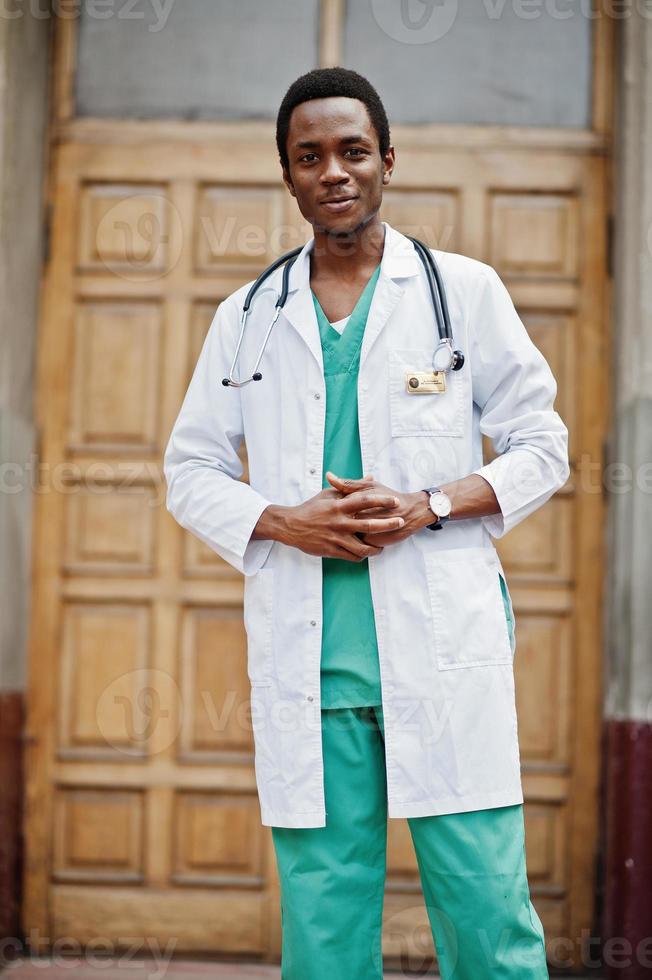 Stylish african american doctor with stethoscope and lab coat posed against door of hospital. photo