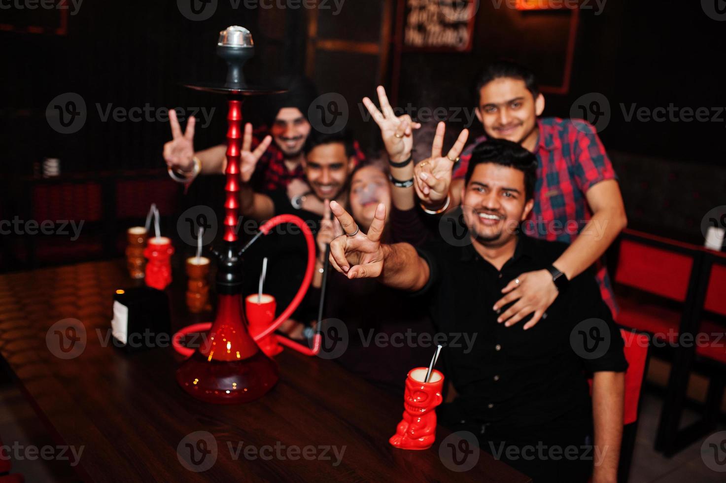 grupo de amigos indios divirtiéndose y descansando en el club nocturno, bebiendo cócteles y fumando narguile. manos con dos dedos hacia arriba. foto