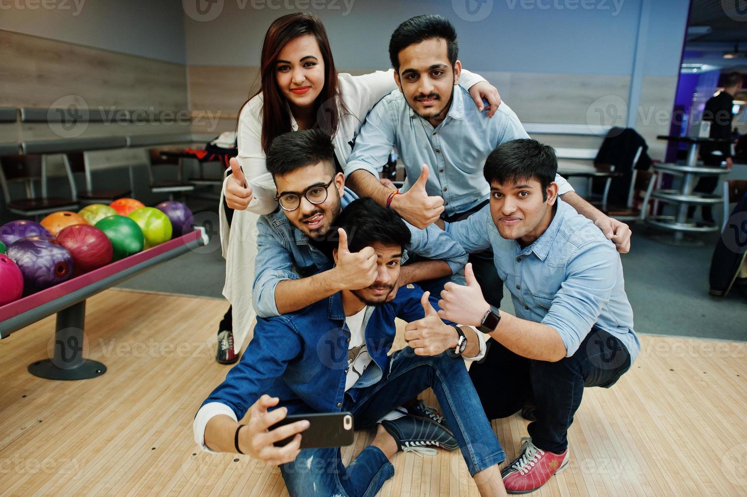 grupo de cinco pueblos del sur de asia descansando y divirtiéndose en el club de bolos. haciendo selfie por teléfono y mostrando el pulgar hacia arriba. foto