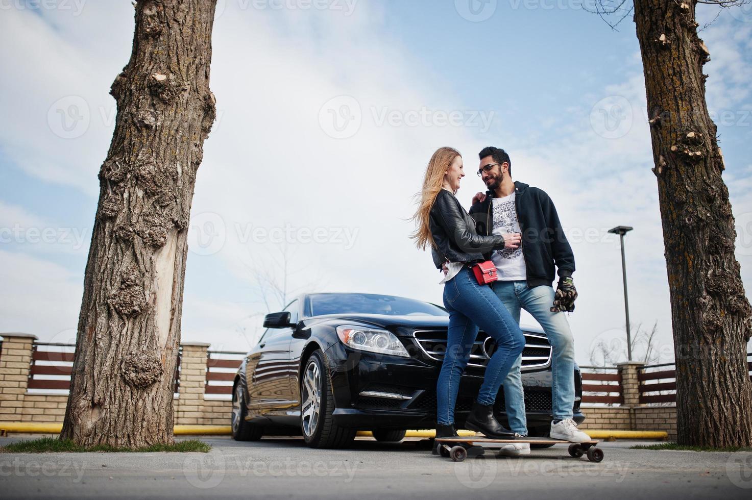 Cool multiracial couple hugs together with longboard against black elegant sport car. photo