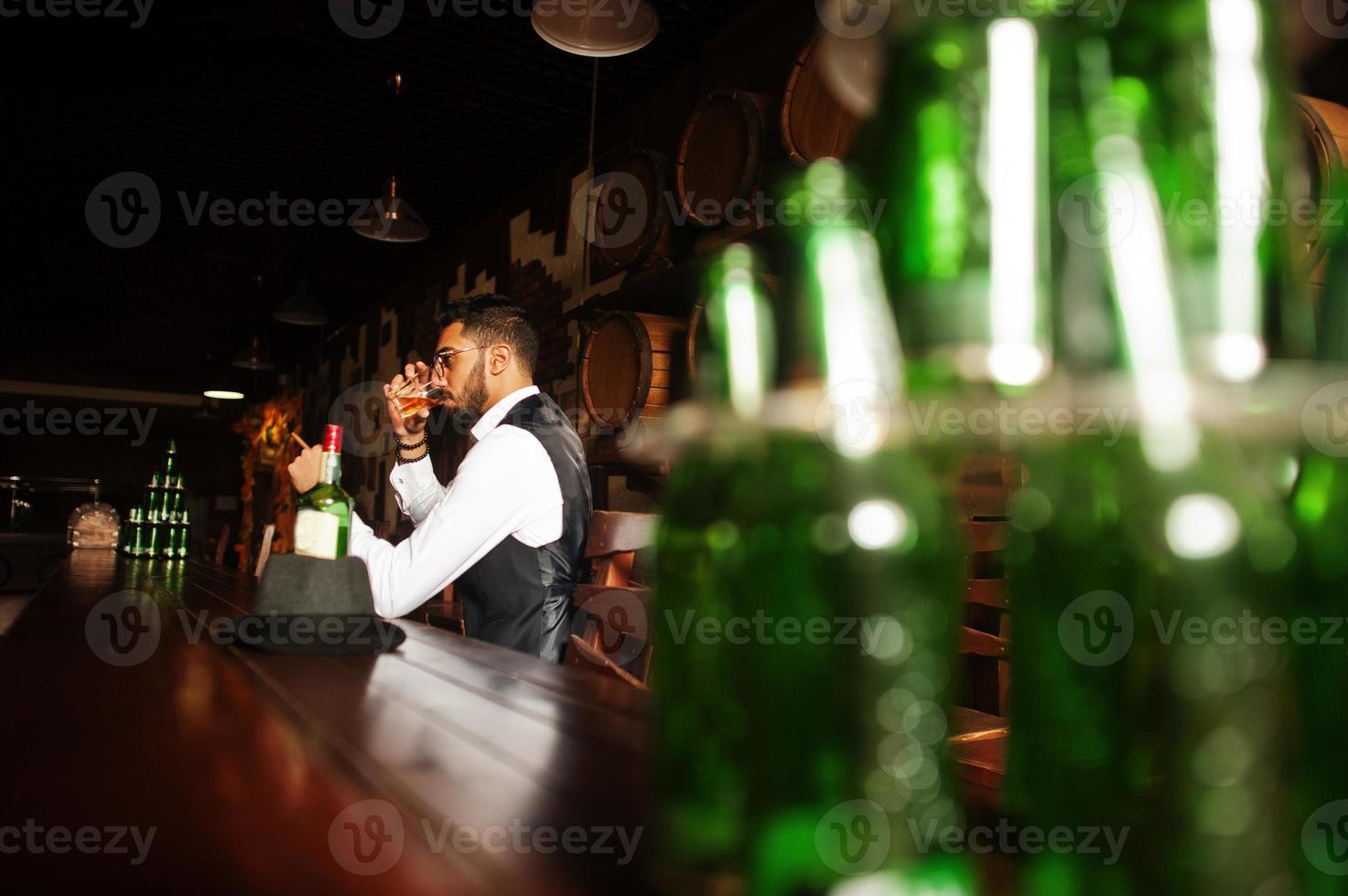 apuesto hombre árabe bien vestido con vaso de whisky y cigarro posado en el pub. foto