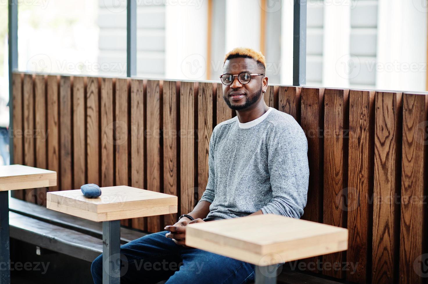 Elegante chico afroamericano con suéter gris y gafas posadas en la calle sentado en una mesa de café de madera. chico negro de moda. foto