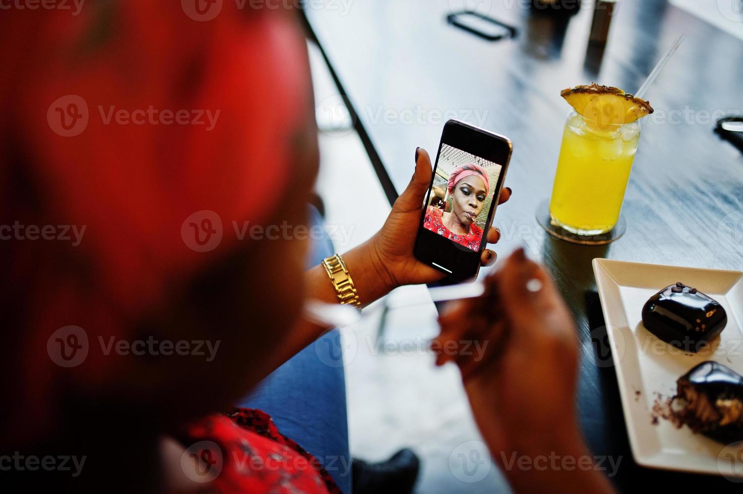 Stylish african woman in red shirt and hat posed indoor cafe, eat chocolate dessert cakes and making selfie at mobile phone. photo