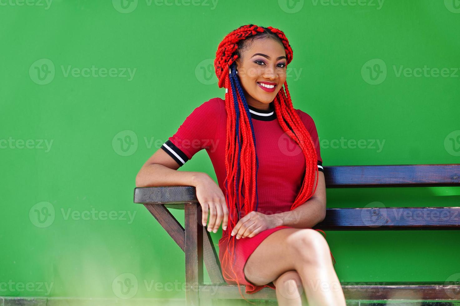 Cute and slim african american girl in red dress with dreadlocks posed on bench background green wall. Stylish black model. photo