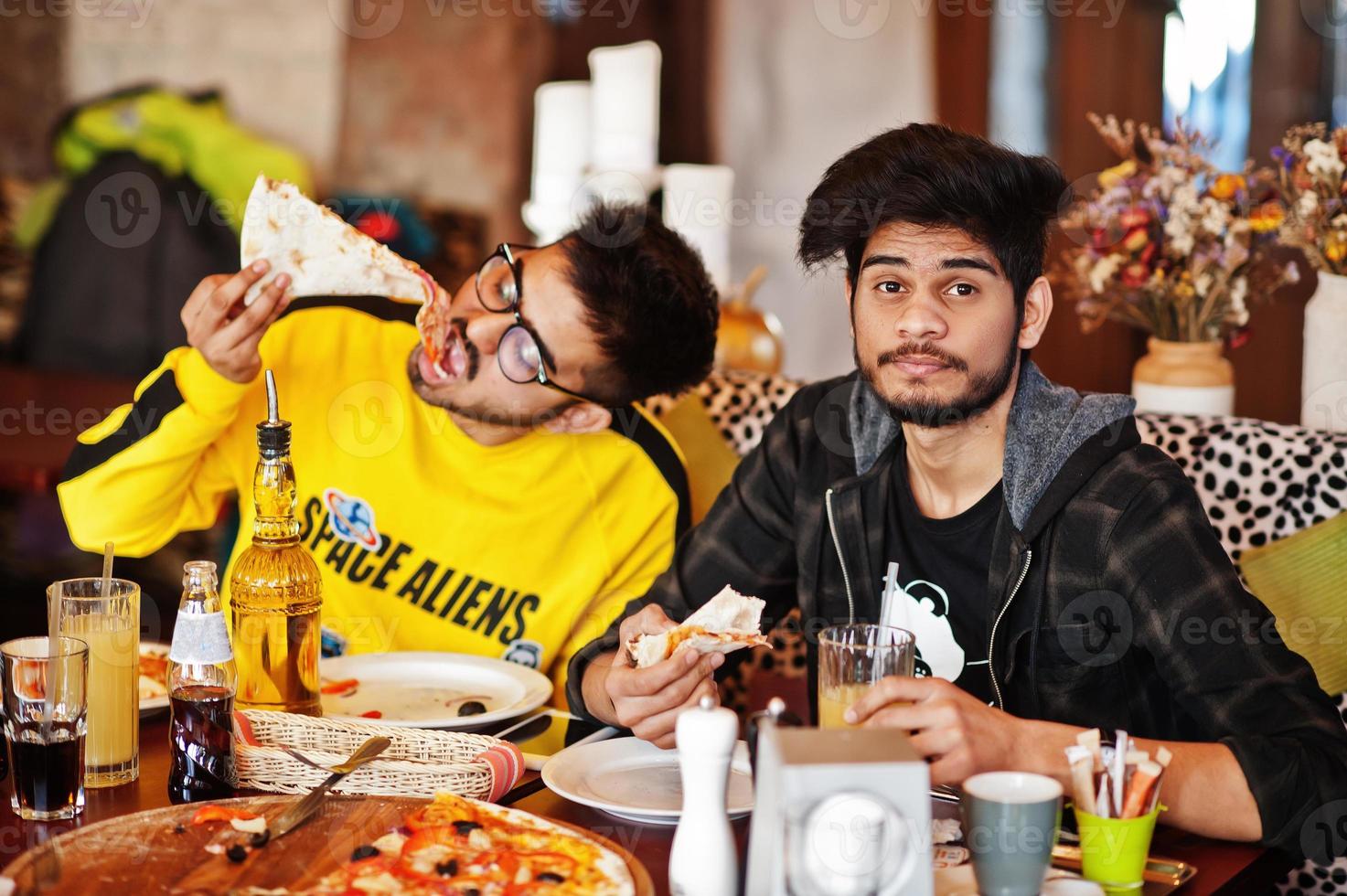 Asian friends guys eating pizza during party at pizzeria. Happy indian people having fun together, eating italian food and sitting on couch. photo