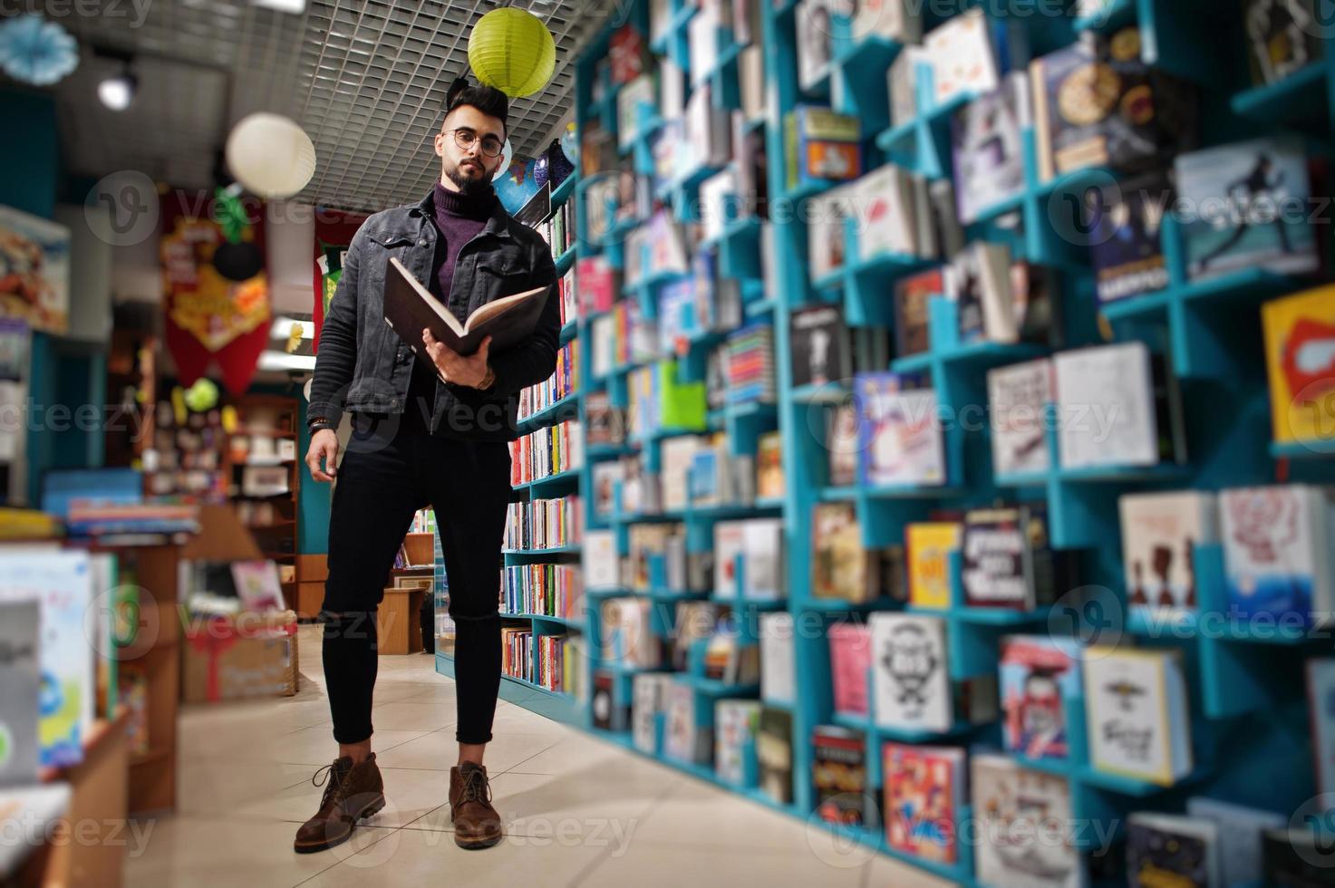 Tall smart arab student man, wear on black jeans jacket and eyeglasses, at library with book at hands. photo