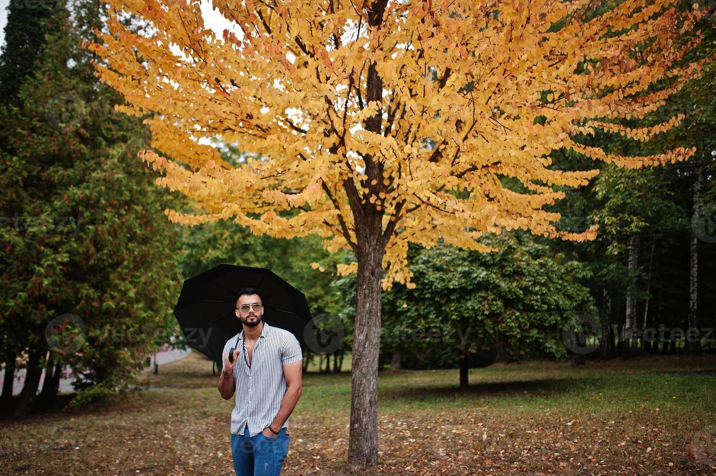 Fashionable tall arab beard man wear on shirt, jeans and sunglasses posed on autumn park with umbrella against yellow leaves tree. photo
