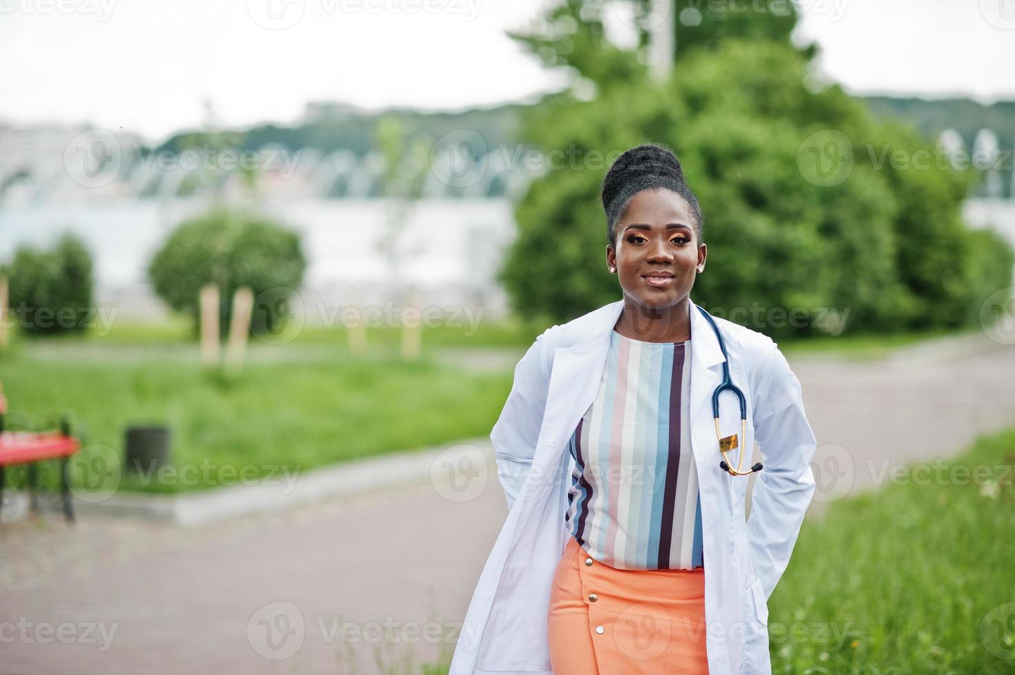 joven doctora afroamericana en bata blanca con un estetoscopio posado al aire libre. foto