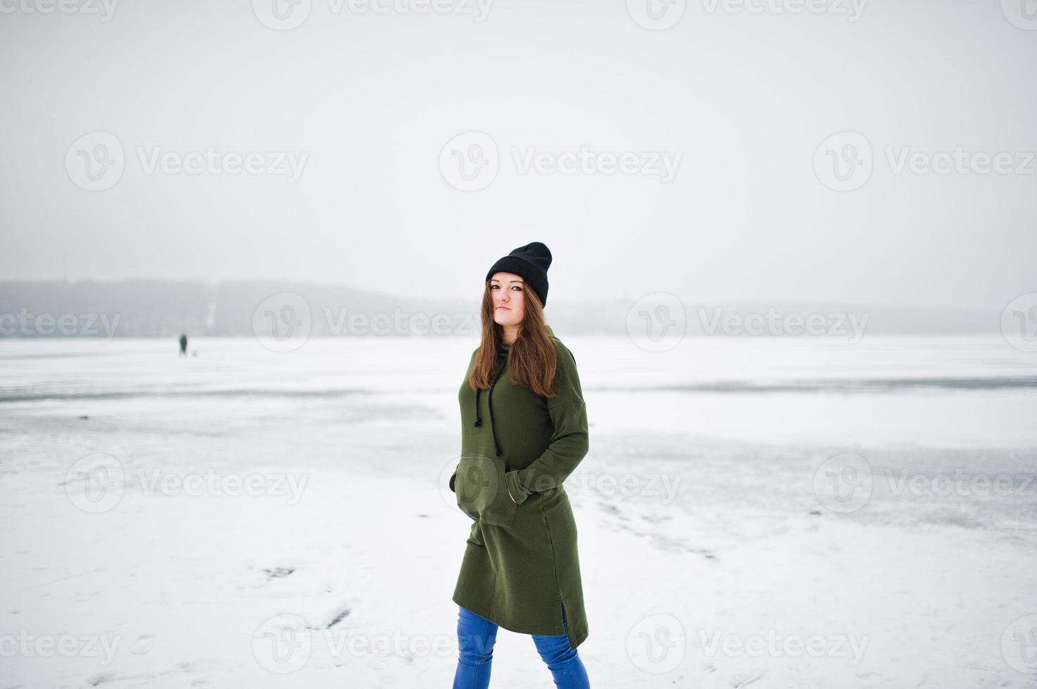 Funny girl wear on long green sweatshirt, jeans and black headwear, at frozen lake in winter day. photo