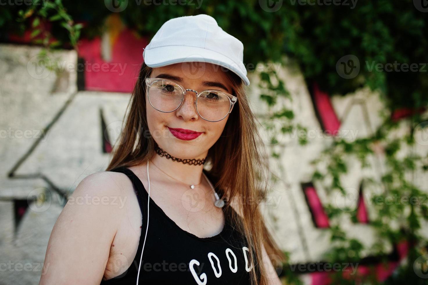Stylish casual hipster girl in cap, glasses and jeans wear, listening music from headphones of mobile phone against large graffiti wall. photo