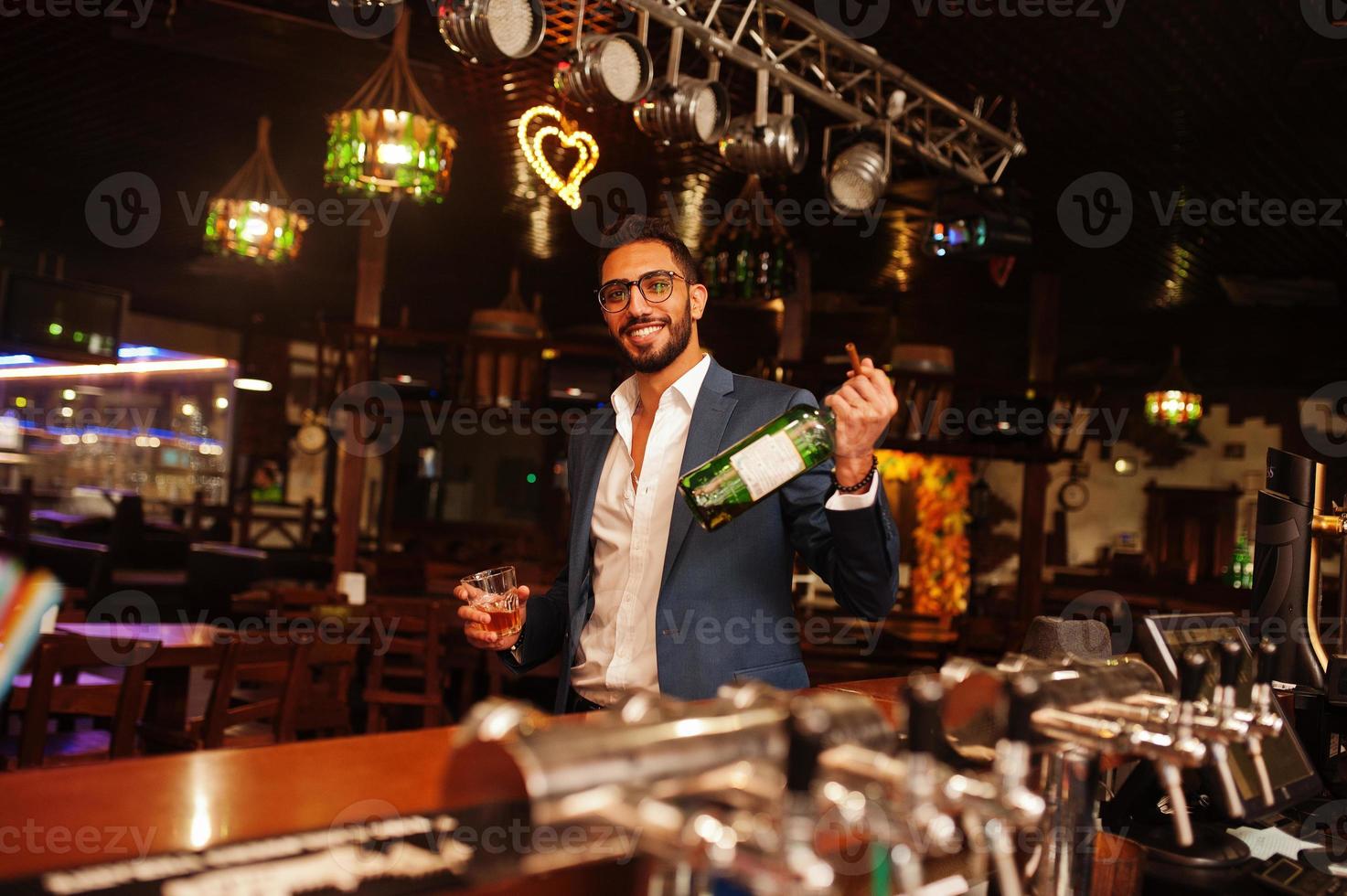 Handsome well-dressed arabian man with glass of whiskey and cigar posed at pub. photo