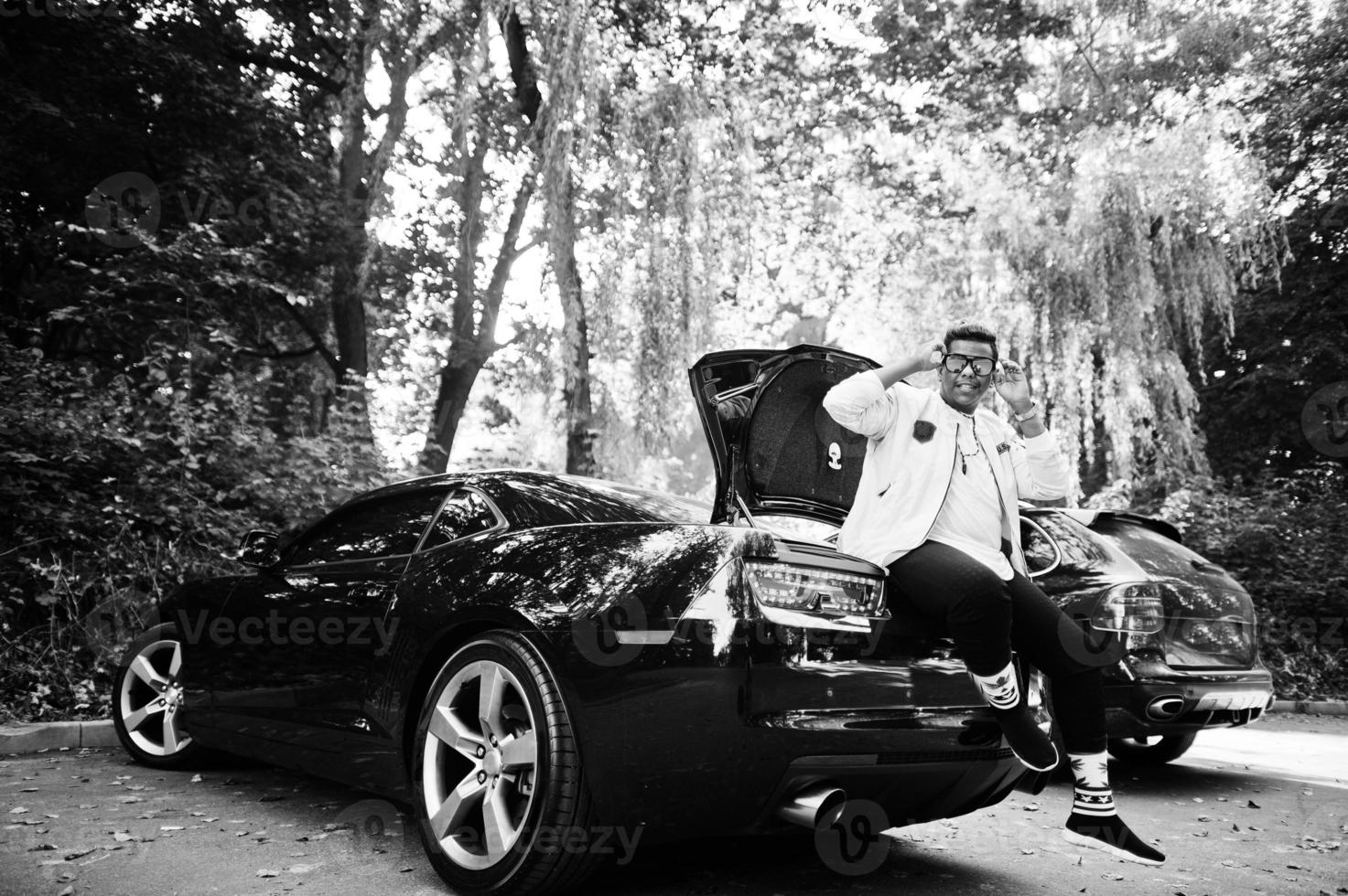 Stylish hipster arab man guy in sunglasses posed outdoor in street and sitting on the trunk his black muscle car. Rich black man. photo