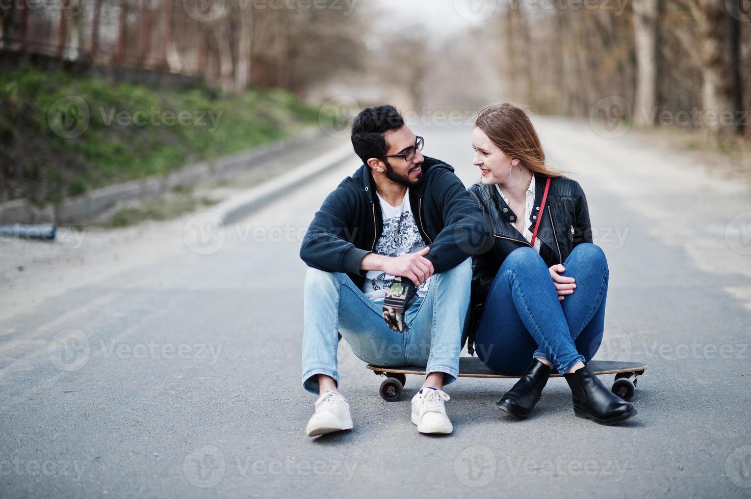 Cool multiracial couple sitting on longboard at road. photo