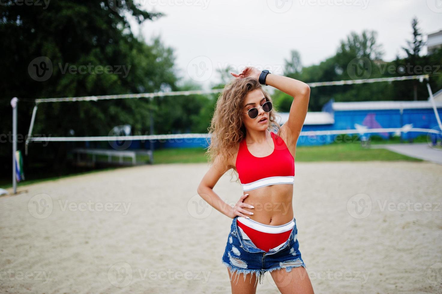 chica sexy modelo rizada en top rojo, pantalones cortos de mezclilla, copa  y gafas de sol posadas en el campo de voleibol de playa. 10415045 Foto de  stock en Vecteezy