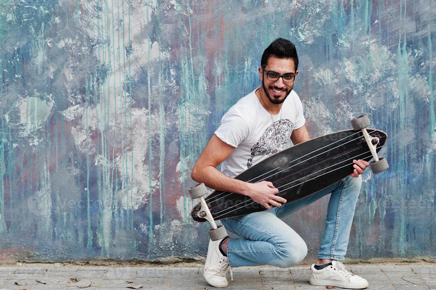 Street style arab man in eyeglasses with longboard posed against colored wall, like he play guitar. photo