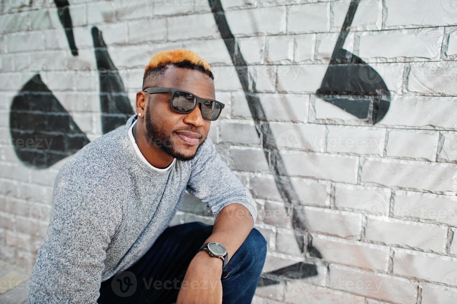 elegante chico afroamericano con suéter gris y gafas de sol negras posadas en la calle. chico negro de moda contra la pared de graffity. foto