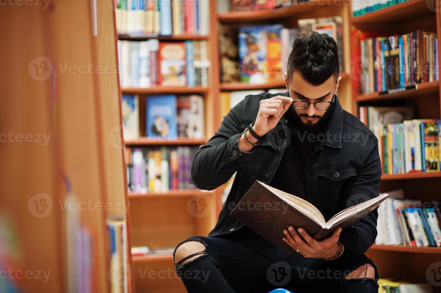 Tall smart arab student man, wear on black jeans jacket and eyeglasses, sitting at library and read book at hands. photo