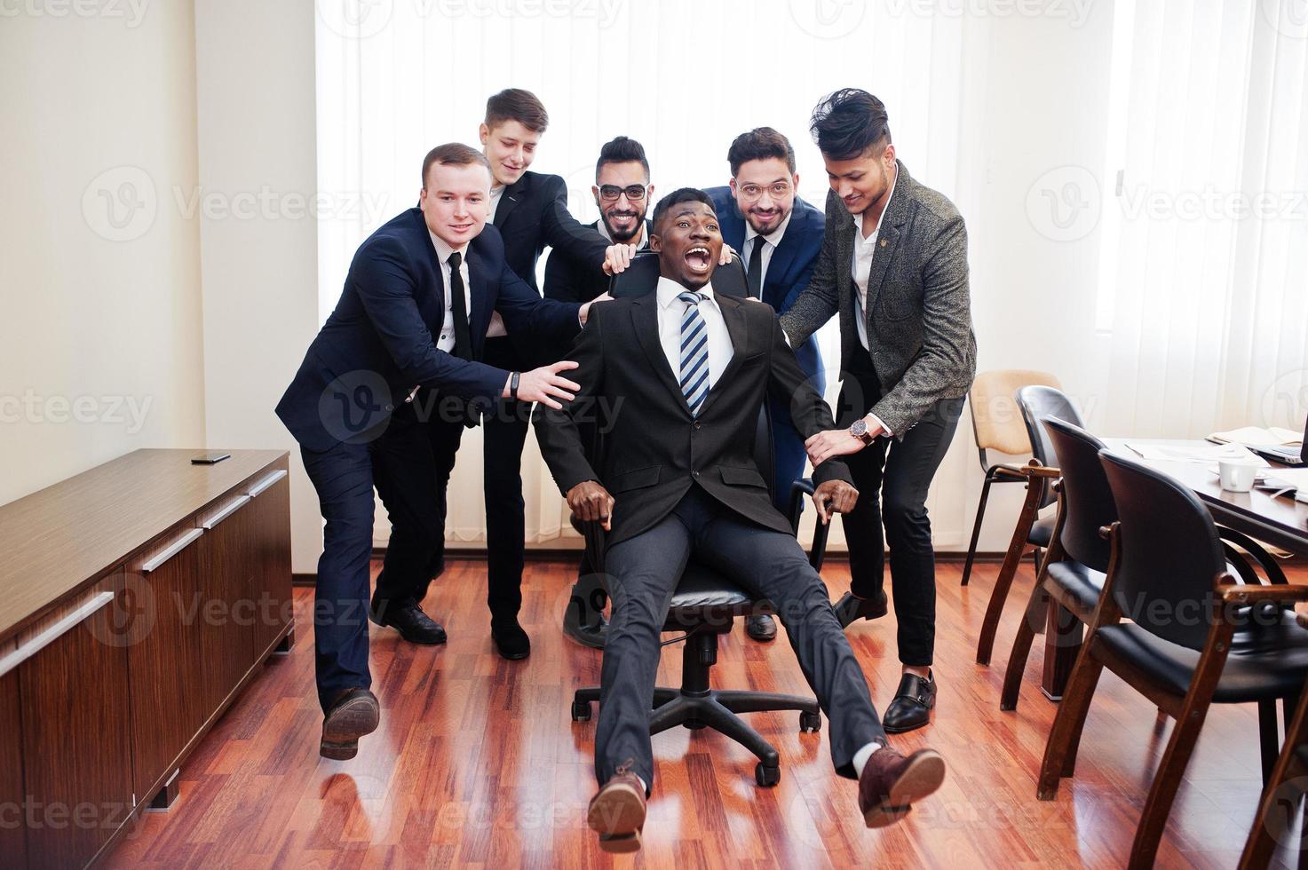 Six multiracial business mans standing at office and roll man on chair. Diverse group of male employees in formal wear having fun. photo