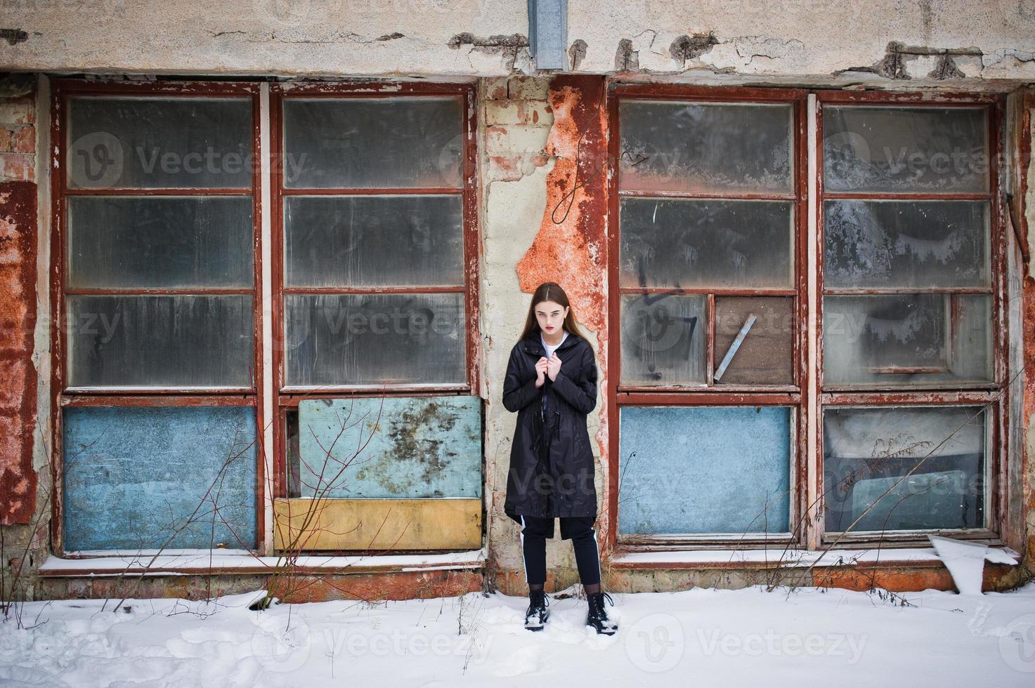 Fashionable long legs brunette model in long black cloak posed outdoor at winter day against old grunge wall with broken windows. photo