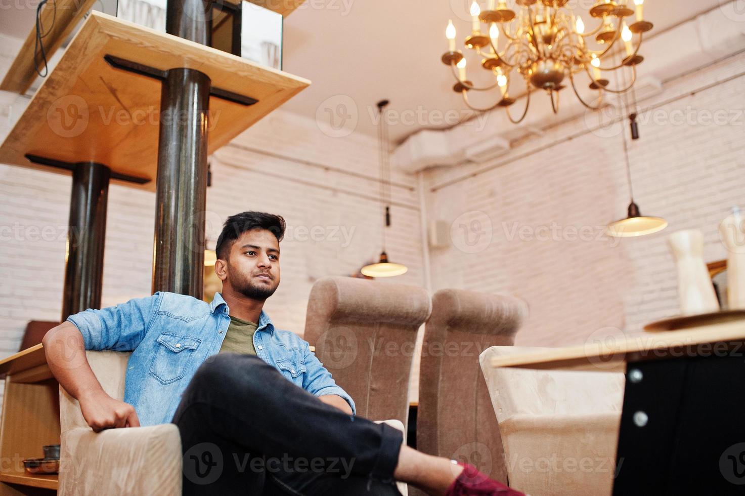 Portrait of handsome successful bearded south asian, young indian freelancer in blue jeans shirt sitting in cafe and having a rest. photo