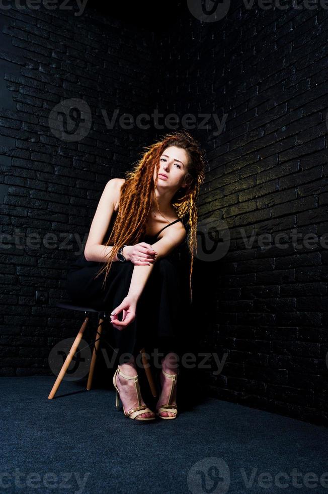 sesión de estudio de una chica de negro en una silla con rastas sobre fondo de ladrillo. foto