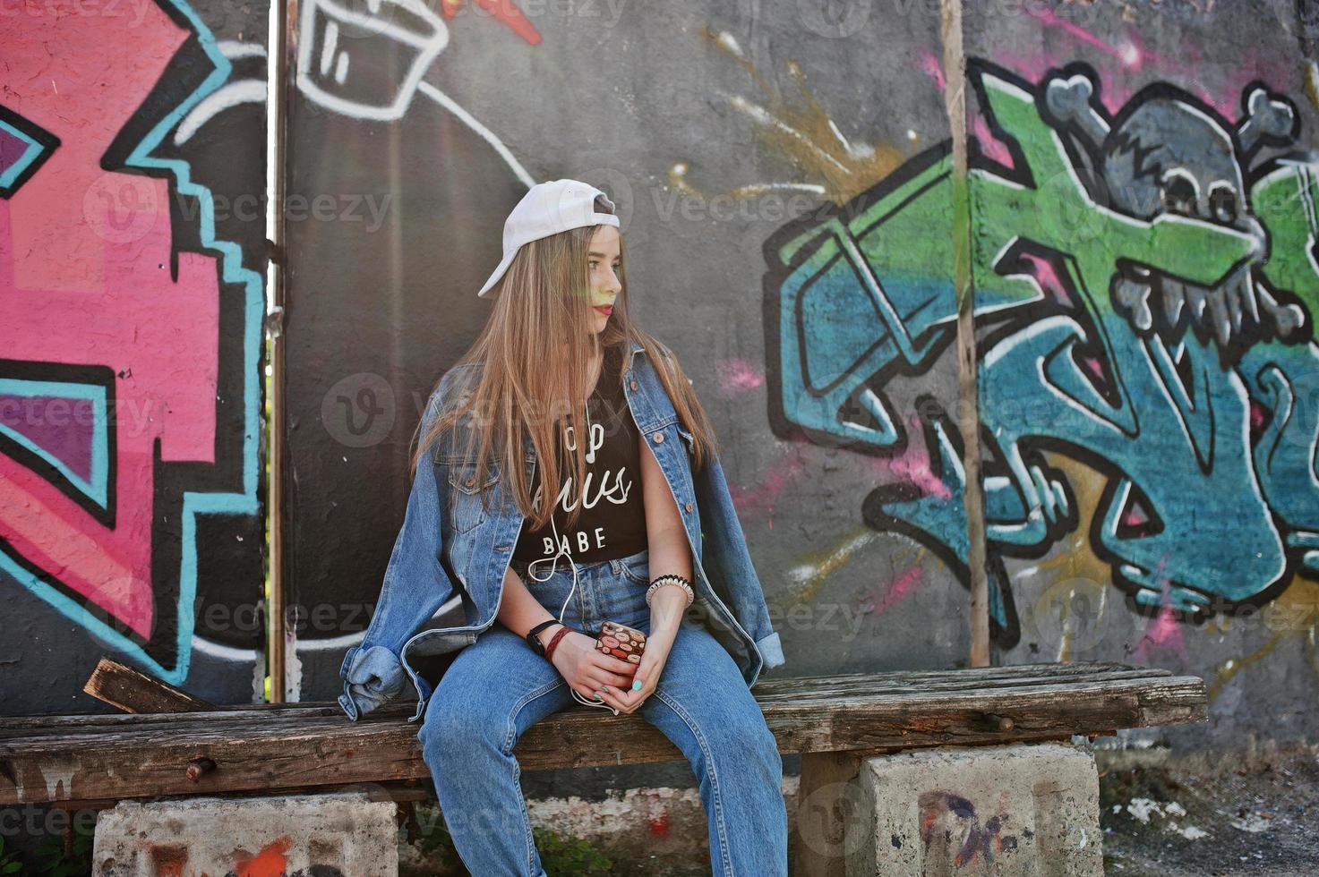 Stylish casual hipster girl in cap and jeans wear listening music from headphones of mobile phone against large graffiti wall with bomb. photo