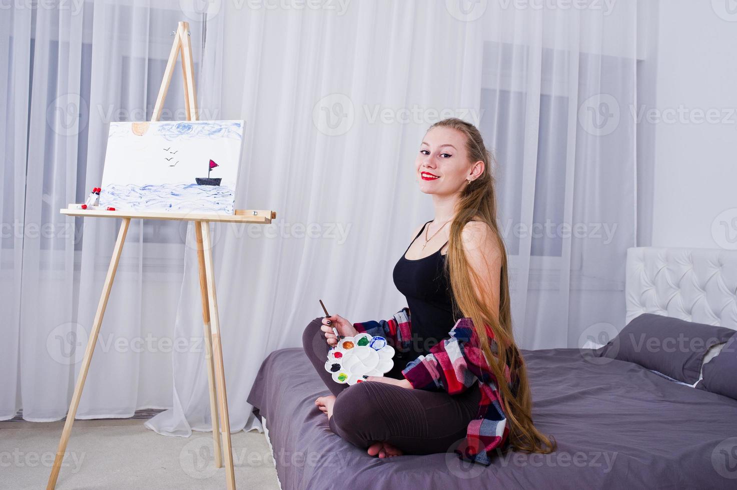 Beautiful woman artist painter with brushes and oil canvas posing bed at room. photo